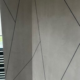 RemontyRs - Dekoracyjna ściana wewnętrzna z szarą, teksturowaną powierzchnią i czarnymi, geometrycznymi liniami, widok z narożnika pomieszczenia, fragment drewnianej podłogi.