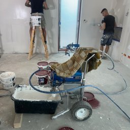 RemontyRs - Dwóch mężczyzn wyrównuje ściany w nowym budynku, jeden na drabinie, drugi na podłodze, z użyciem szpachli i agregatu malarskiego przykrytego kocem.