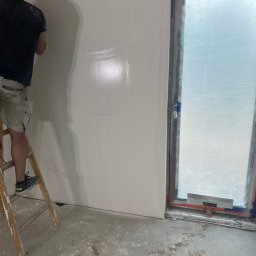 RemontyRs - Mężczyzna na drabinie wygładza ścianę w pomieszczeniu z oknem zabezpieczonym folią, widoczne ślady zaprawy na podłodze i drabinie.