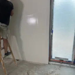 Mężczyzna na drabinie wygładza ścianę w pomieszczeniu z oknem zabezpieczonym folią, widoczne ślady zaprawy na podłodze i drabinie.