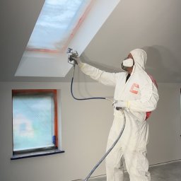 RemontyRs - Pracownik w kombinezonie ochronnym maluje natryskowo sufit z oknem dachowym, okno i framuga okna zabezpieczone folią, widoczne ślady farby na podłodze.