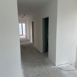 RemontyRs - Korytarz w trakcie remontu: świeżo pomalowane białe ściany, surowy beton na podłodze, widoczne kable i gniazdka elektryczne, w tle drzwi z surową ościeżnicą i widoczną konstrukcją.