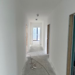 RemontyRs - Korytarz w trakcie prac wykończeniowych: świeżo pomalowane ściany na biało, widoczne kable i przedłużacz na podłodze, ościeżnice drzwi bez zamontowanych drzwi.