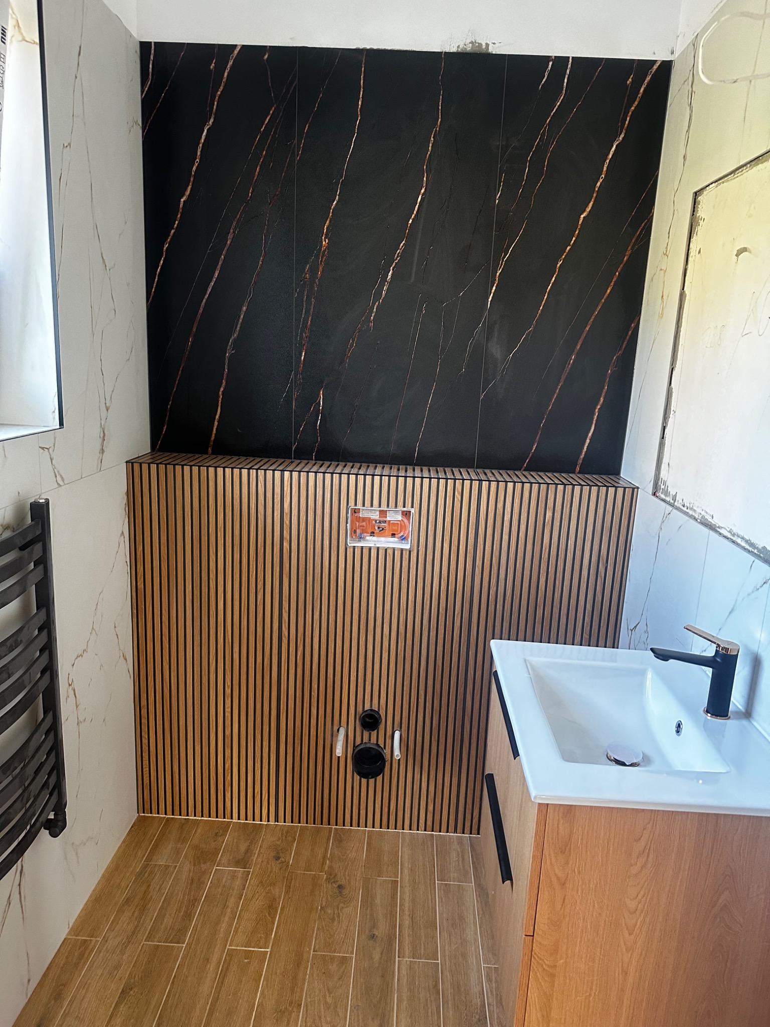 Nowoczesna łazienka z czarnymi płytkami imitującymi marmur ze złotymi żyłami, drewnianą boazerią lamelową ukrywającą stelaż WC, oraz umywalką na szafce z drewnopodobnym fornirem i czarną armaturą.