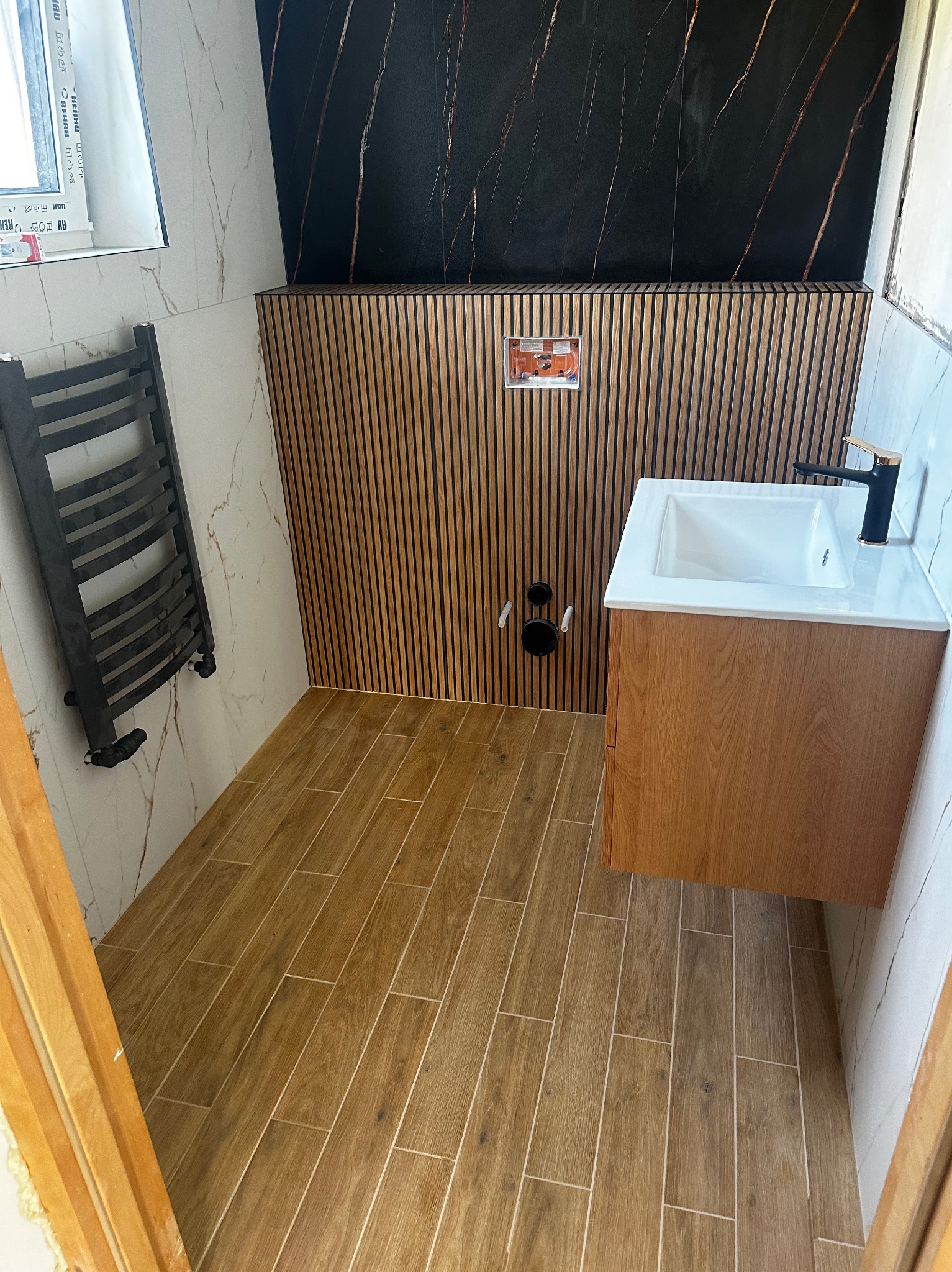 Nowoczesna łazienka z drewnianą zabudową umywalki, grzejnikiem drabinkowym w kolorze grafitowym i płytkami imitującymi drewno na podłodze, uzupełniona czarną ścianą z marmurowym wzorem.