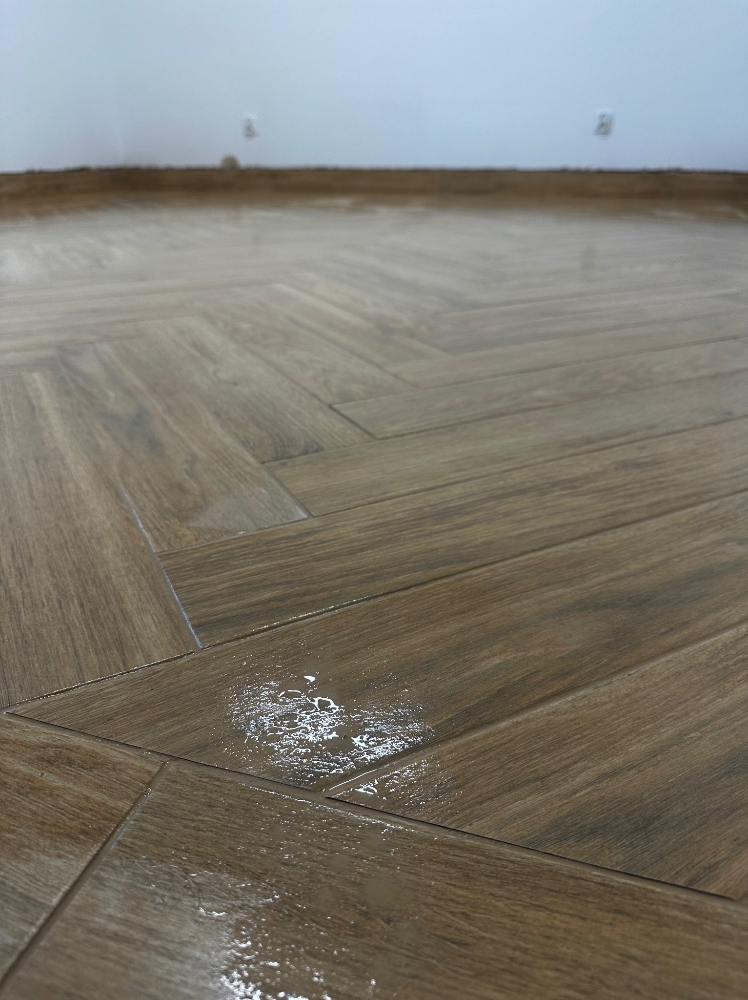 Ułożona w jodełkę podłoga z paneli imitujących drewno, z widocznymi śladami świeżego kleju lub wody na powierzchni, ściana w tle przygotowana do malowania.