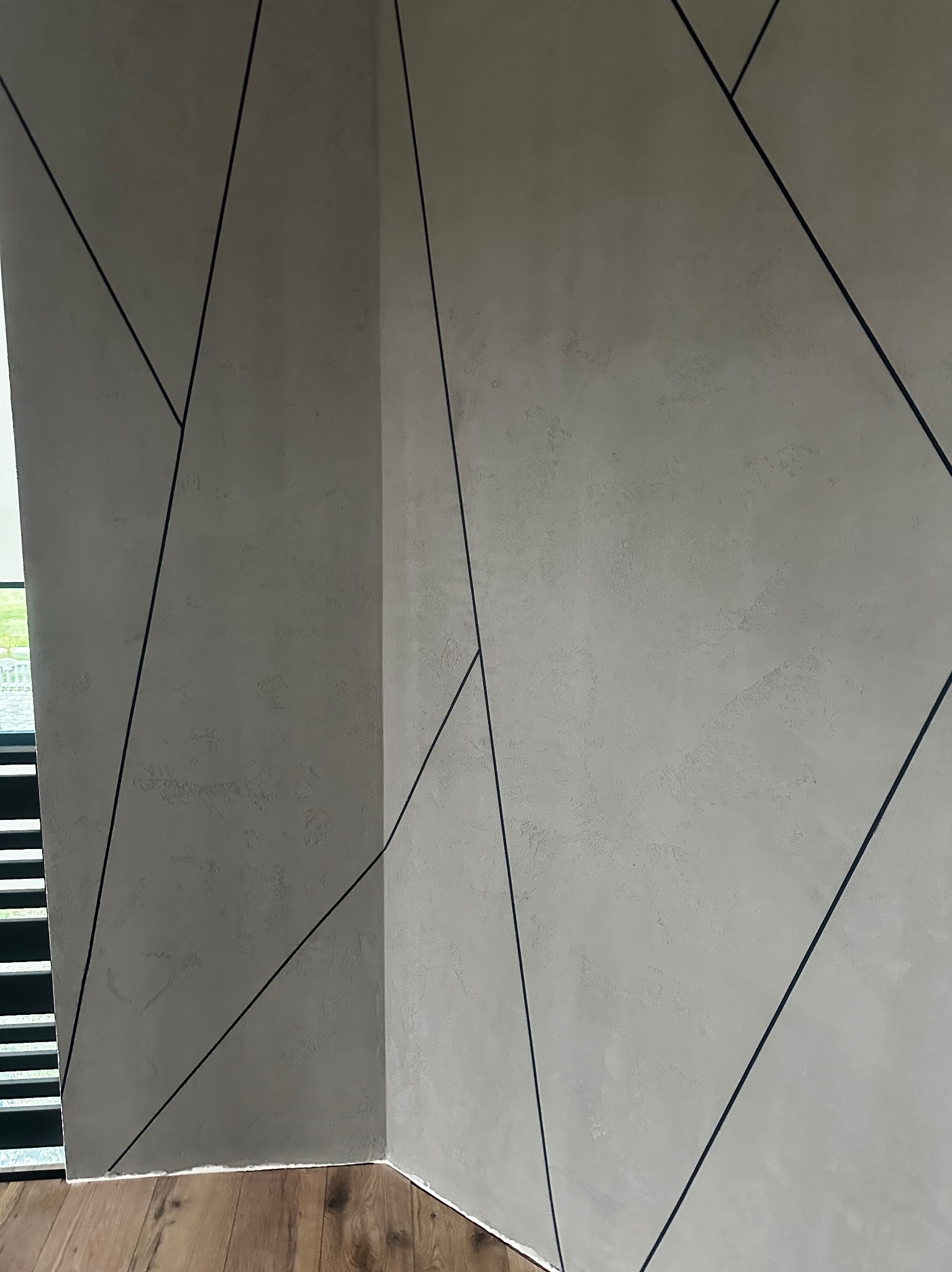 Dekoracyjna ściana wewnętrzna z szarą, teksturowaną powierzchnią i czarnymi, geometrycznymi liniami, widok z narożnika pomieszczenia, fragment drewnianej podłogi.
