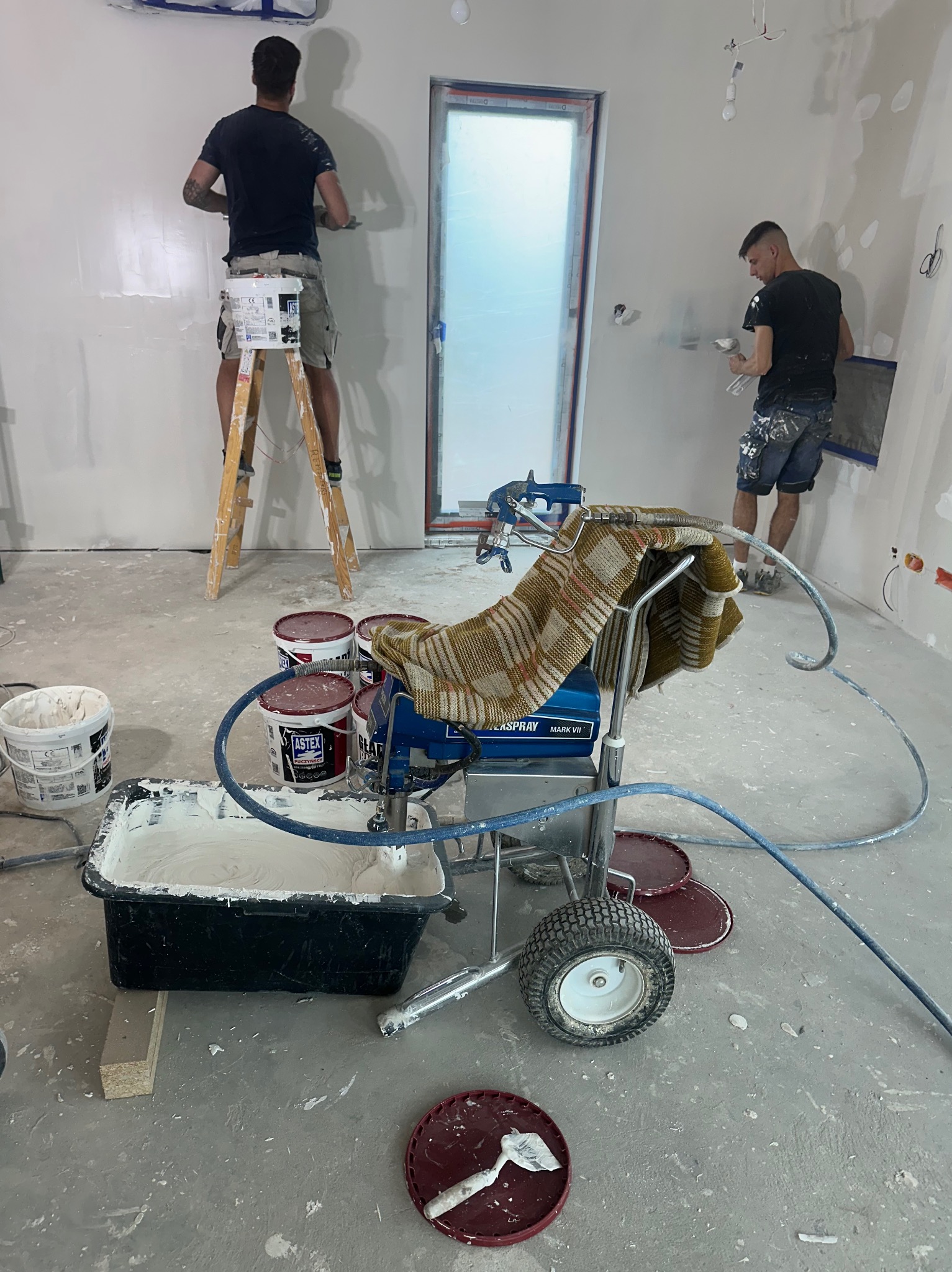 Dwóch mężczyzn wyrównuje ściany w nowym budynku, jeden na drabinie, drugi na podłodze, z użyciem szpachli i agregatu malarskiego przykrytego kocem.