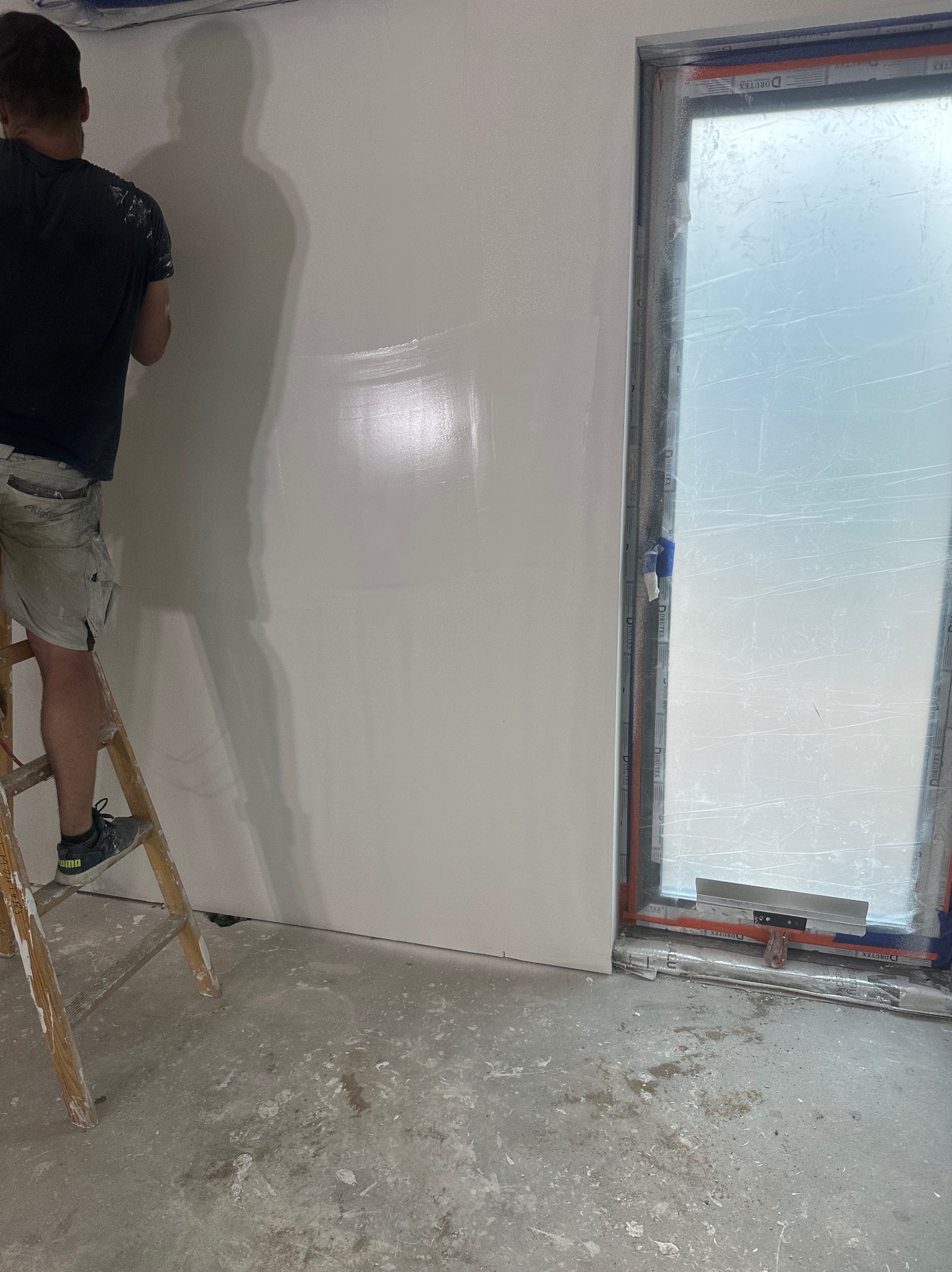 Mężczyzna na drabinie wygładza ścianę w pomieszczeniu z oknem zabezpieczonym folią, widoczne ślady zaprawy na podłodze i drabinie.