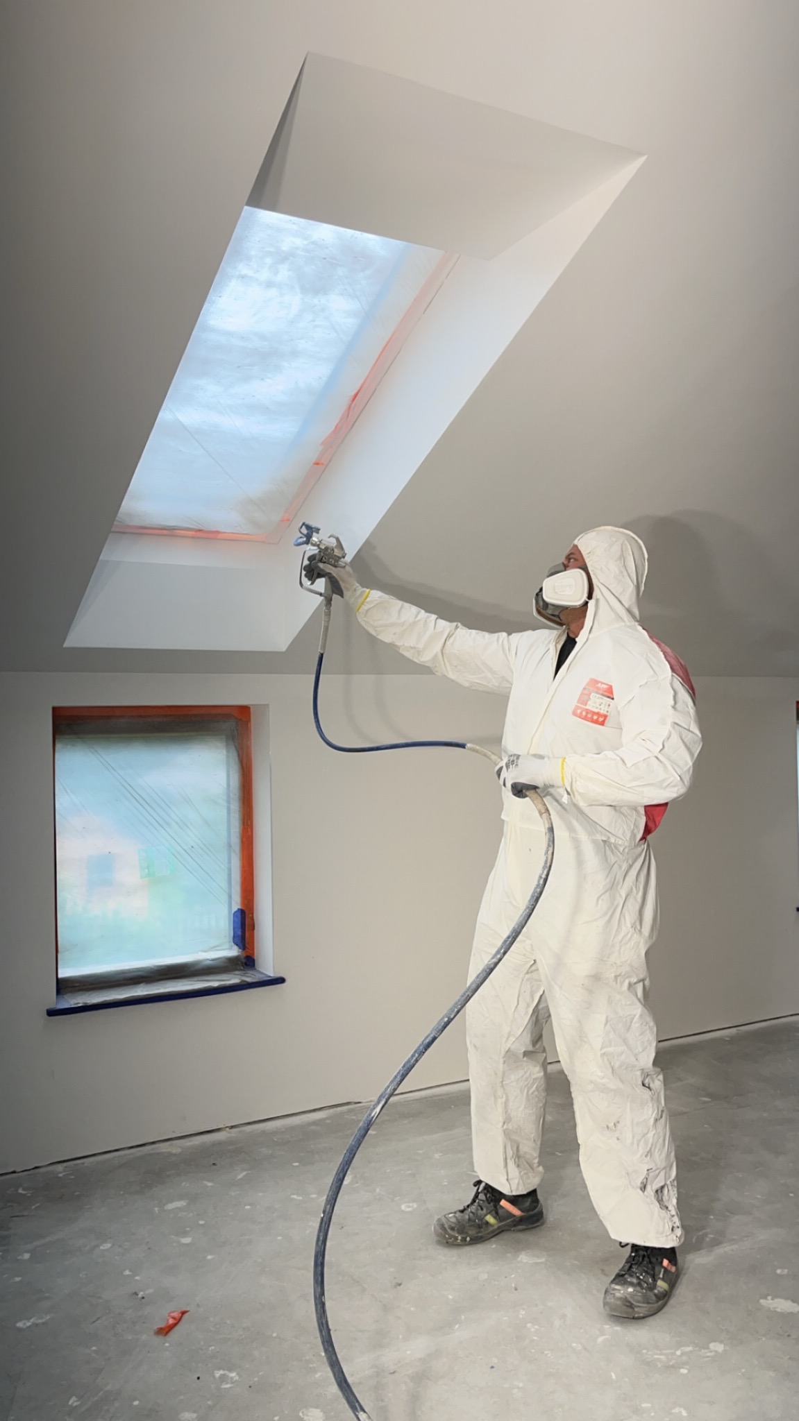 Pracownik w kombinezonie ochronnym maluje natryskowo sufit z oknem dachowym, okno i framuga okna zabezpieczone folią, widoczne ślady farby na podłodze.