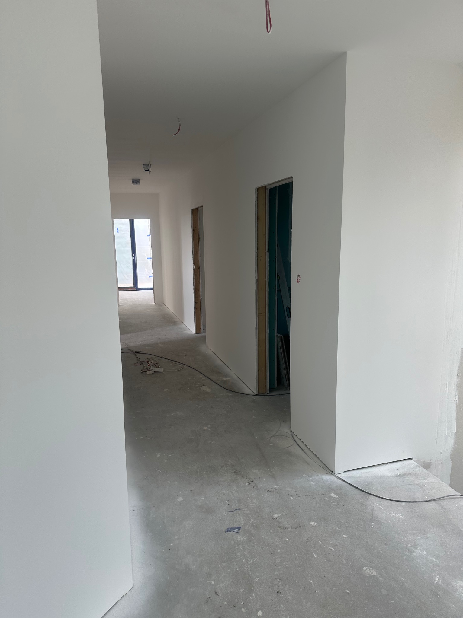 Korytarz w trakcie remontu: świeżo pomalowane białe ściany, surowy beton na podłodze, widoczne kable i gniazdka elektryczne, w tle drzwi z surową ościeżnicą i widoczną konstrukcją.