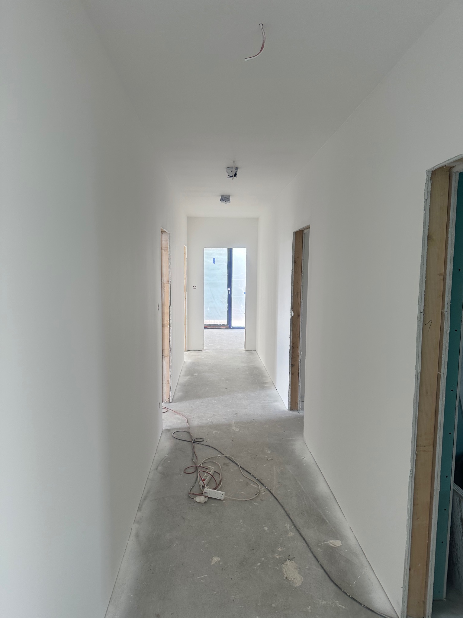 Korytarz w trakcie prac wykończeniowych: świeżo pomalowane ściany na biało, widoczne kable i przedłużacz na podłodze, ościeżnice drzwi bez zamontowanych drzwi.