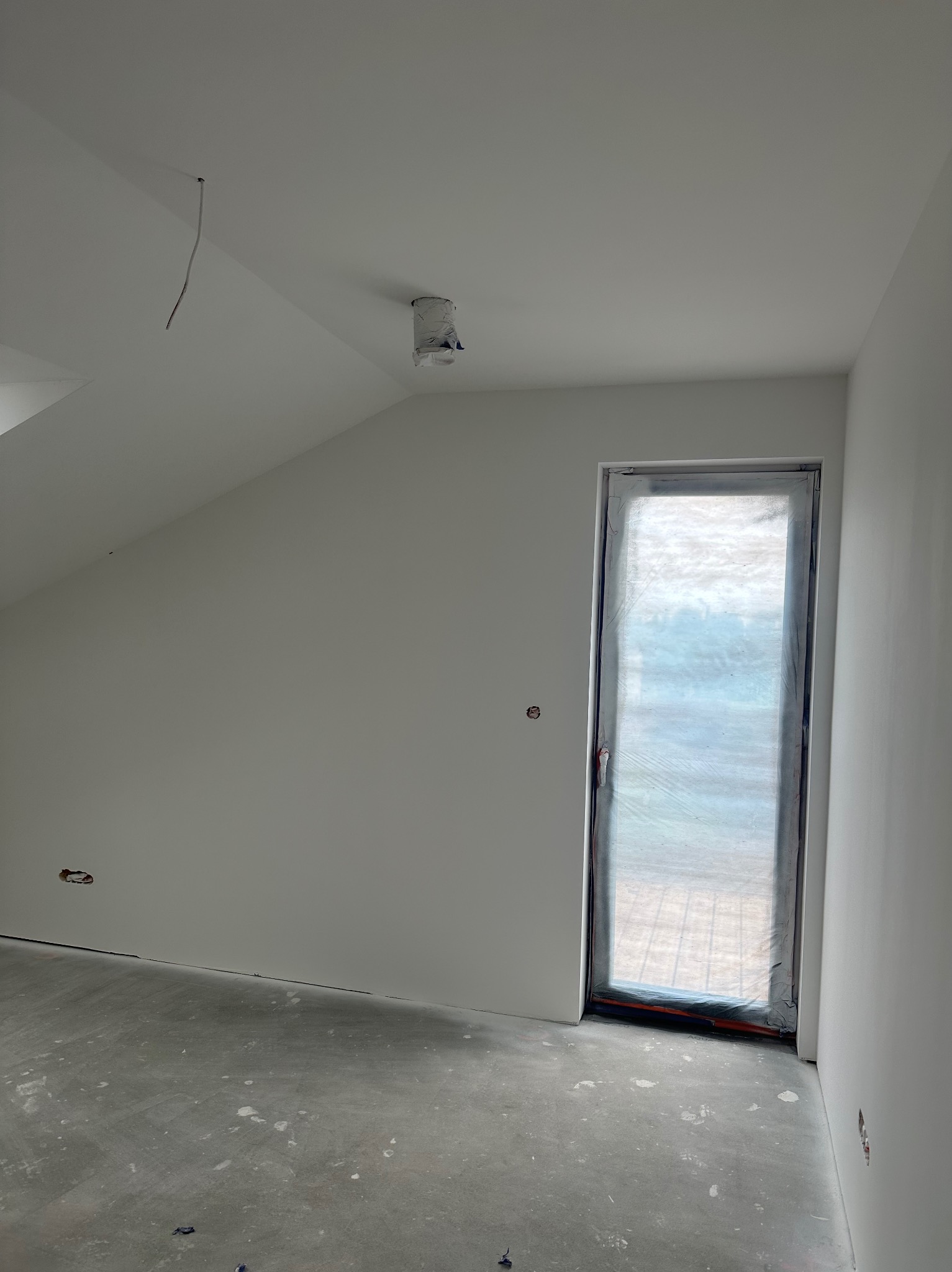 Wnętrze pokoju w trakcie prac wykończeniowych, ściany pokryte świeżą, białą warstwą, okno zabezpieczone folią ochronną, widoczne wystające przewody elektryczne.