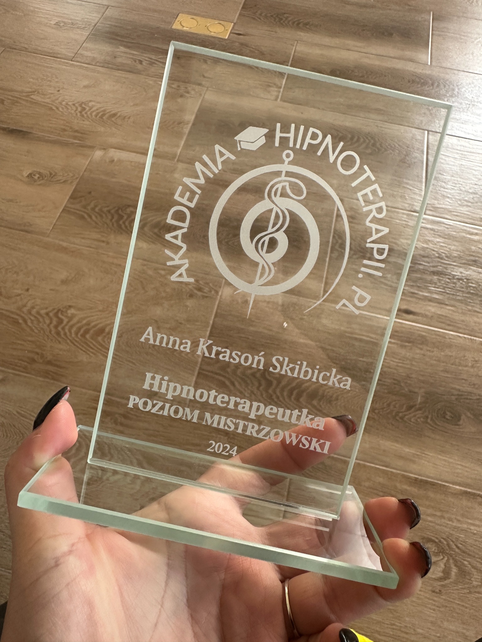 Przezroczysta statuetka z wygrawerowanym napisem 'Akademia Hipnoterapii.pl', symbolem laski Eskulapa, imieniem i nazwiskiem 'Anna Krasoń Skibicka' oraz tytułem 'Hipnoterapeutka POZIOM MISTRZOWSKI...
