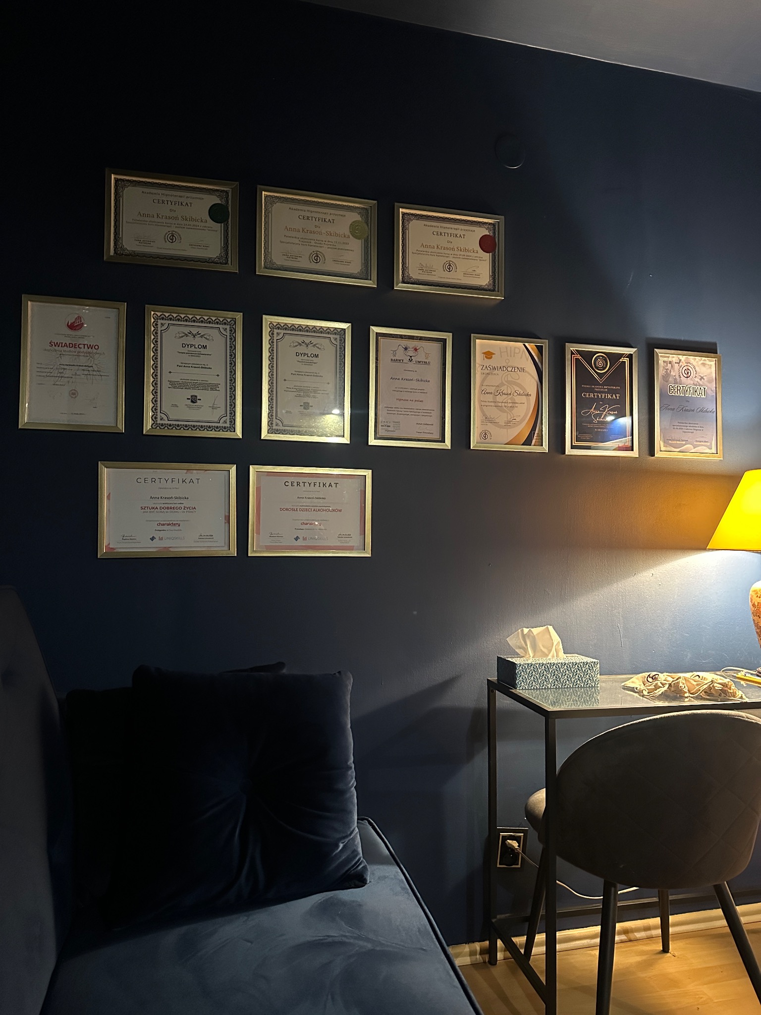 Ściana gabinetu z dyplomami i certyfikatami w złotych ramkach, biurko z lampką i chusteczkami higienicznymi, fragment sofy w odcieniach granatu.