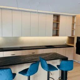 Nowoczesna kuchnia z drewnianymi szafkami, czarnym blatem i wyspą z trzema turkusowymi hokerami, oświetlenie LED pod szafkami wiszącymi i na suficie.