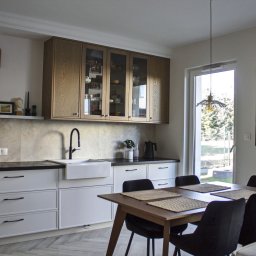 Dopasowany Studio - Jasna kuchnia z drewnianymi szafkami wiszącymi z przeszklonymi frontami, białymi szafkami dolnymi z czarnymi uchwytami, czarnym kranem i ceramicznym zlewem. Na blacie czajnik i mała roślina. W tle...