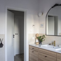 Dopasowany Studio - Nowoczesna łazienka z drewnianą szafką pod umywalką, złotą baterią, okrągłym lustrem z ozdobnym wykończeniem, otwartymi drzwiami i wzorzystą podłogą w tle.