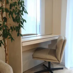 Toaletka z lustrem wbudowana w szafę, tapicerowane krzesło na czarnej podstawie, roślina doniczkowa, jasne drewniane podłogi i białe zasłony w tle.