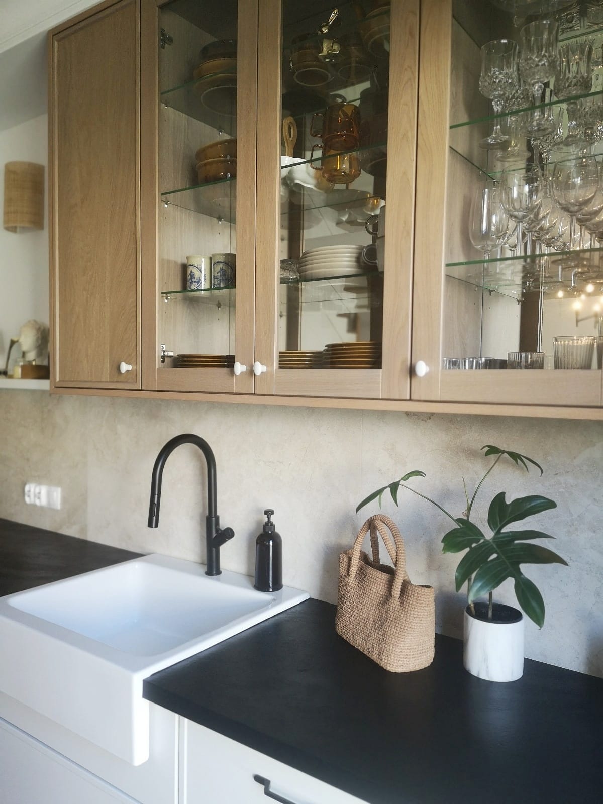 Jasna kuchnia z drewnianymi szafkami z przeszklonymi frontami, białym zlewem farmerskim i czarną baterią oraz blatem, uzupełniona o wiklinową torebkę i roślinę doniczkową.