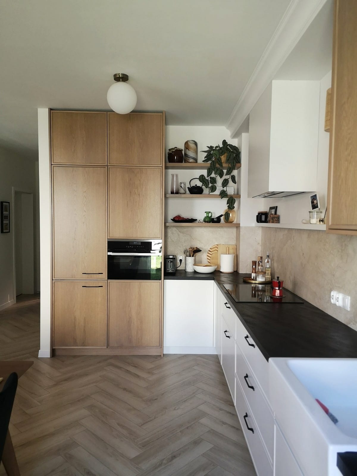 Nowoczesna kuchnia z drewnianymi szafkami, czarnym blatem i białymi frontami szuflad, uzupełniona o otwarte półki z dekoracjami i roślinami, z podłogą ułożoną w jodełkę.