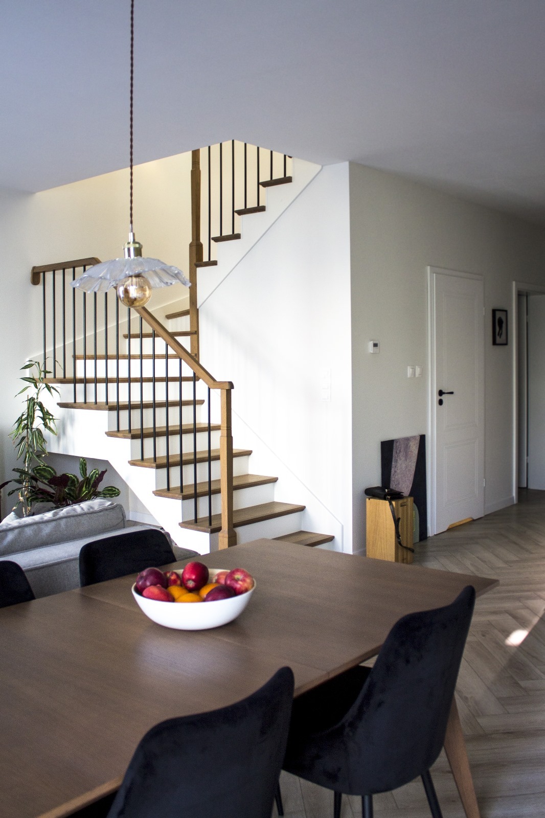 Wnętrze domu z drewnianymi schodami z czarnymi balustradami, lampą wiszącą z kloszem w kształcie kwiatu nad stołem z owocami w białej misie, oraz podłogą ułożoną w jodełkę.