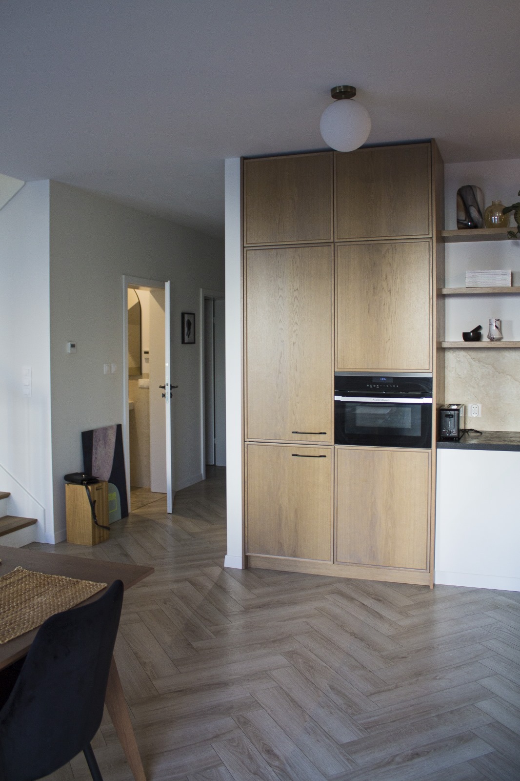 Kuchnia w stylu nowoczesnym z zabudową meblową z forniru dębowego, piekarnikiem w zabudowie, parkietem ułożonym w jodełkę i widokiem na łazienkę w głębi.