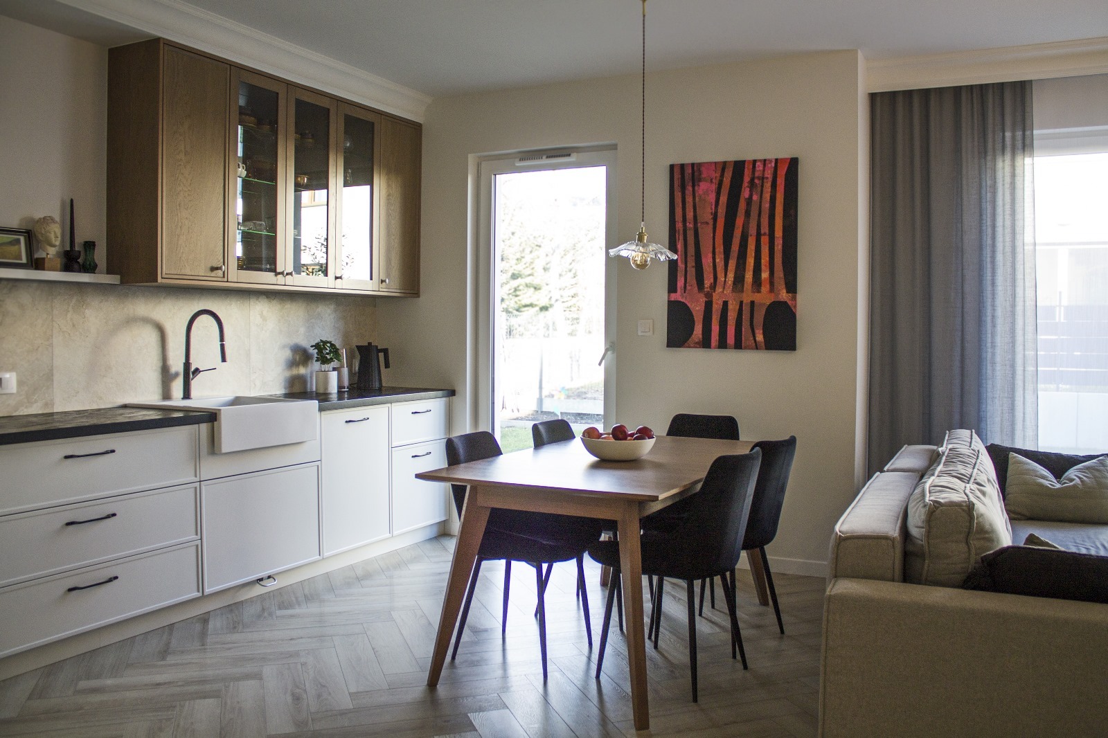 Jasne wnętrze: kuchnia z białymi szafkami, drewnianymi górnymi szafkami z przeszkleniami, stół z czarnymi krzesłami i owocami, szara sofa z poduszkami, parkiet w jodełkę, abstrakcyjny obraz...