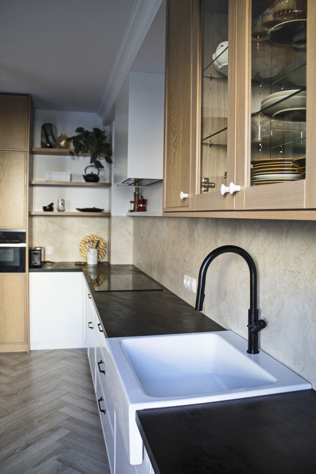 Nowoczesna kuchnia z białymi szafkami, czarnym blatem i armaturą, drewnianymi szafkami wiszącymi z przeszklonymi frontami oraz drewnianą podłogą ułożoną w jodełkę.