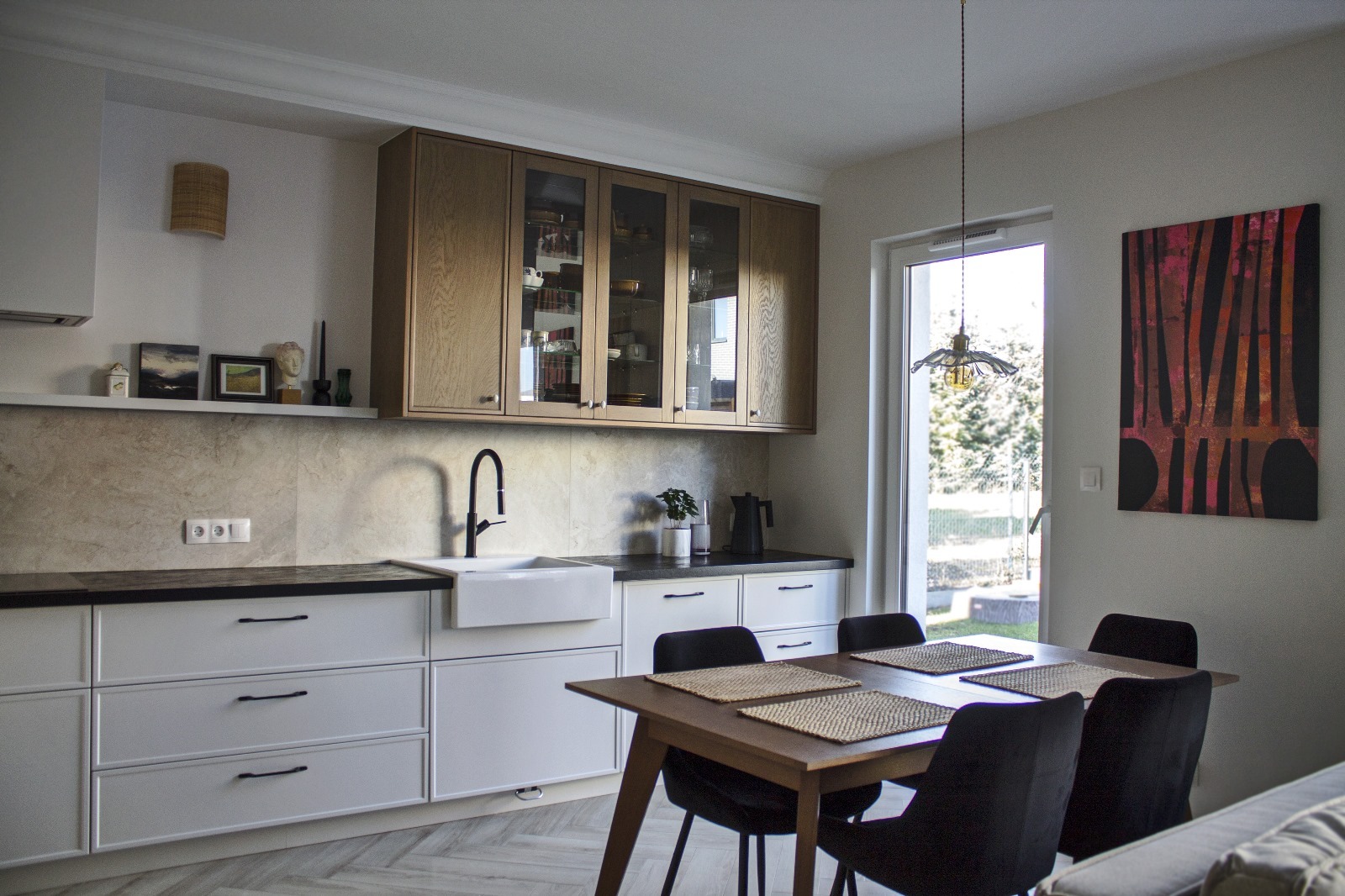 Jasna kuchnia z drewnianymi szafkami wiszącymi z przeszklonymi frontami, białymi szafkami dolnymi z czarnymi uchwytami, czarnym kranem i ceramicznym zlewem. Na blacie czajnik i mała roślina. W tle...