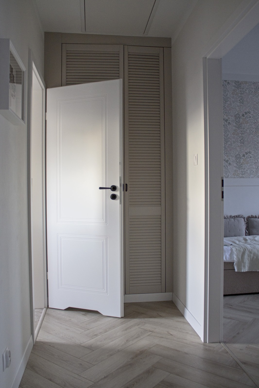 Otwarta biała, płycinowa drzwi wewnętrzne z czarną klamką, przylegająca do szafy z żaluzjowymi drzwiami w odcieniach szarości, widok na sypialnię z tapetą w kwiaty i łóżkiem, podłoga w jodełkę.