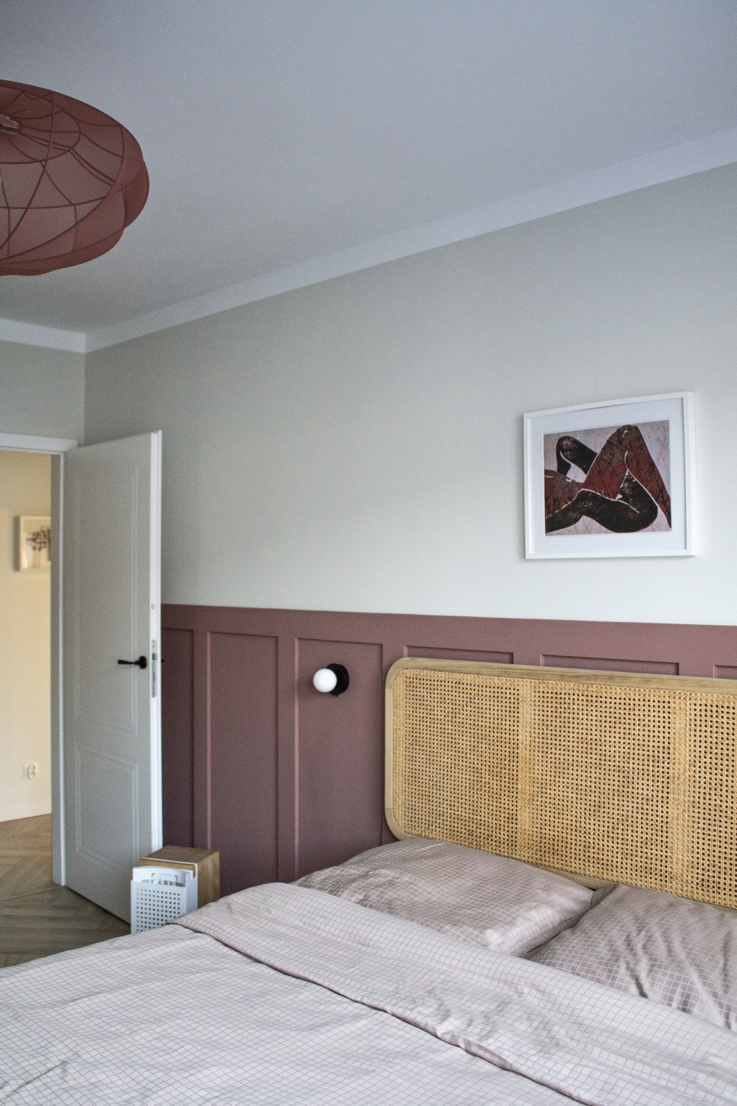 Sypialnia z rattanowym zagłówkiem, panelami ściennymi w kolorze dusty rose, lampką nocną z kulistym kloszem i obrazem w białej ramie nad łóżkiem.