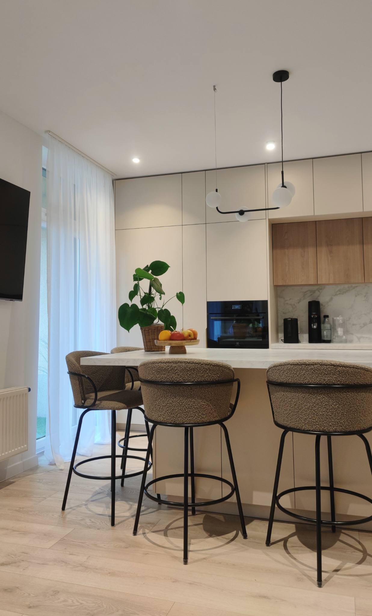 Jasna kuchnia z wyspą, na której stoją trzy krzesła barowe z brązowym obiciem, na blacie wyspy stoi doniczka z rośliną monstera i misa z owocami, nad wyspą wisi nowoczesna lampa.