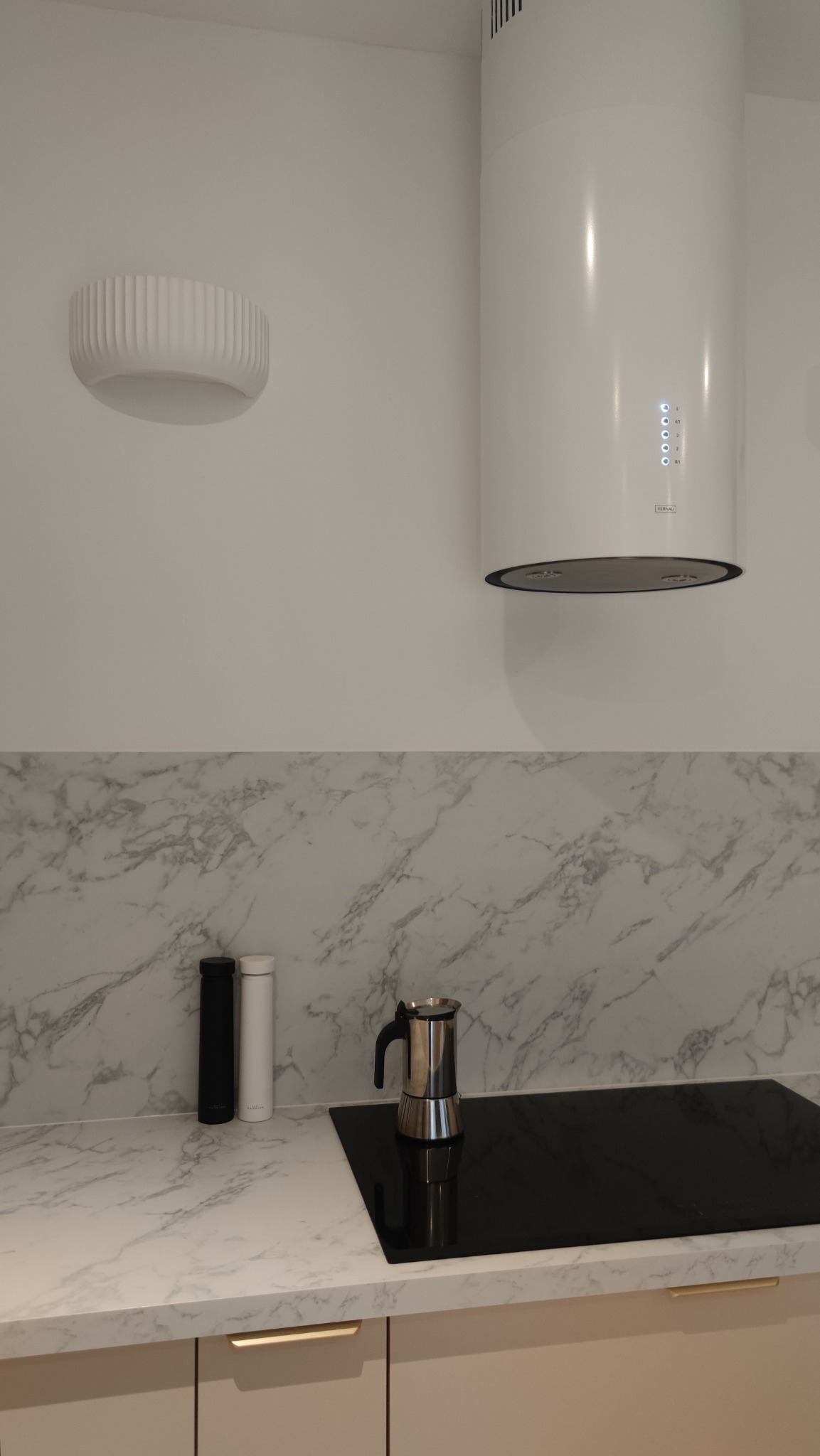 Nowoczesna kuchnia z marmurowym blatem, płytą indukcyjną i srebrną kawiarką, uzupełniona minimalistycznym oświetleniem sufitowym i okrągłym okapem Kernau.