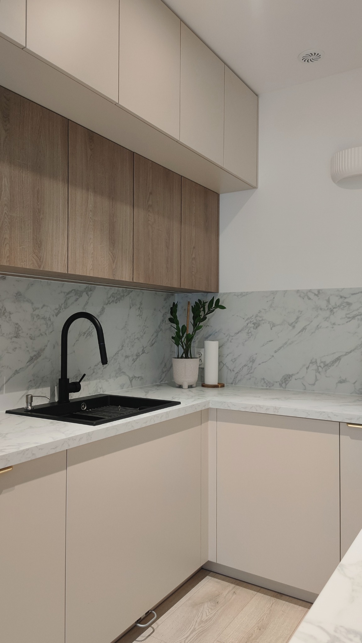 Nowoczesna kuchnia z beżowymi szafkami, drewnianymi górnymi szafkami, marmurowym blatem i czarnym zlewem z baterią, uzupełniona rośliną w ceramicznej doniczce.