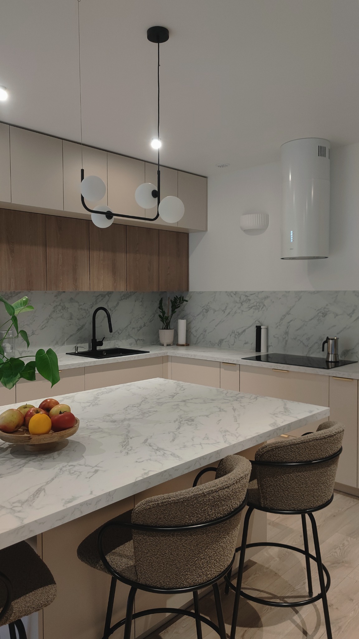 Nowoczesna kuchnia z marmurowym blatem, minimalistycznym oświetleniem i krzesłami barowymi z teksturowaną tapicerką, uzupełniona owocami w drewnianej misie.