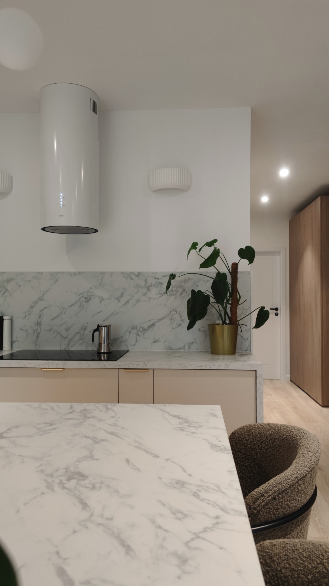 Jasna kuchnia z marmurowym blatem, białym okapem cylindrycznym, płytą indukcyjną, kawiarką i rośliną Monstera w złotej doniczce, widok na drewnianą szafę w korytarzu.