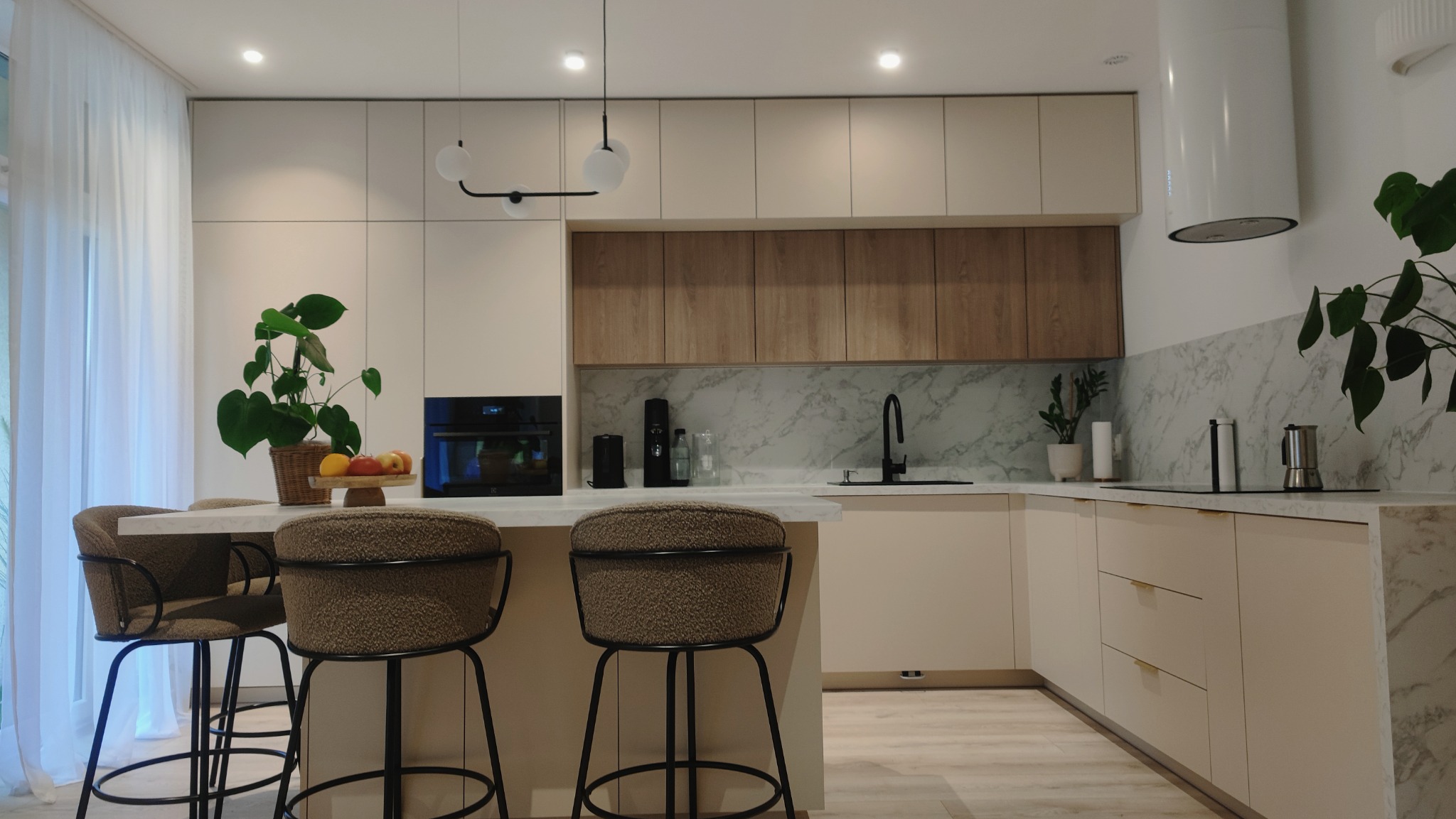 Jasna kuchnia z wyspą, wykończona marmurowym blatem i drewnianymi szafkami, oświetlona nowoczesnymi lampami. Na wyspie stoją dwa krzesła barowe z brązowym siedziskiem i czarnymi nogami, a na blacie...