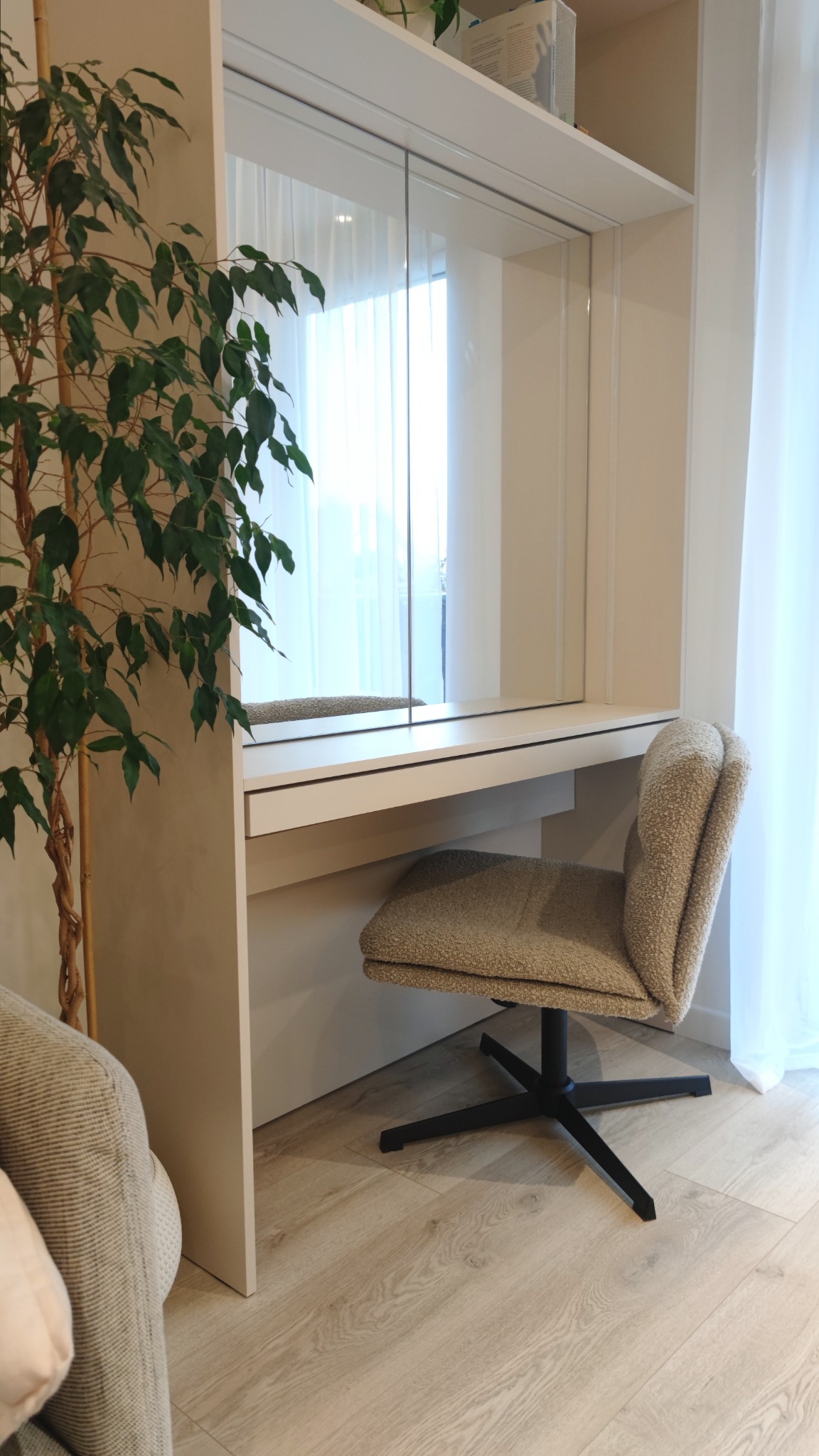 Toaletka z lustrem wbudowana w szafę, tapicerowane krzesło na czarnej podstawie, roślina doniczkowa, jasne drewniane podłogi i białe zasłony w tle.