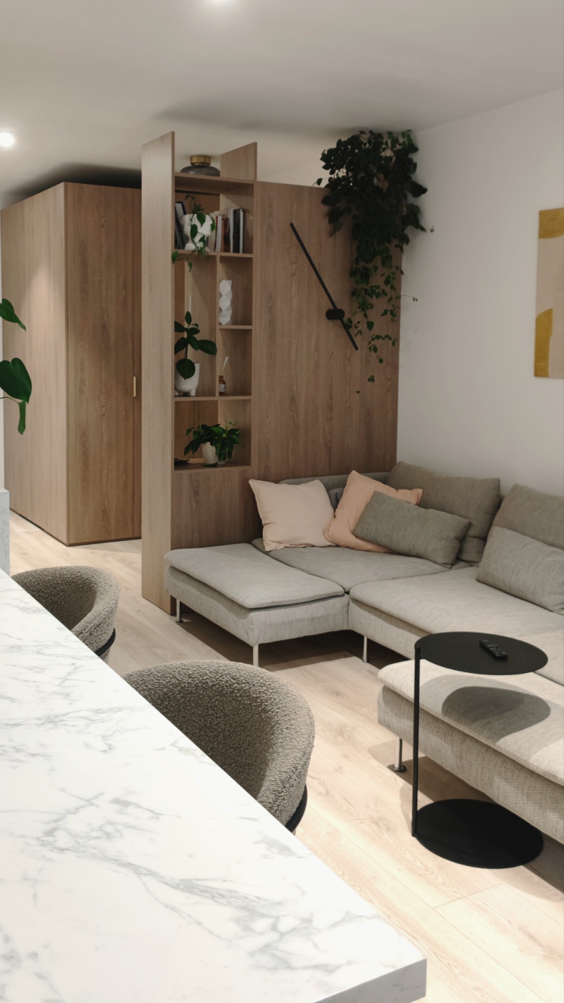 Jasne wnętrze z narożną sofą w odcieniach szarości, marmurowy blat stołu na pierwszym planie, drewniane szafy i regał z roślinami doniczkowymi, duży zegar na ścianie.