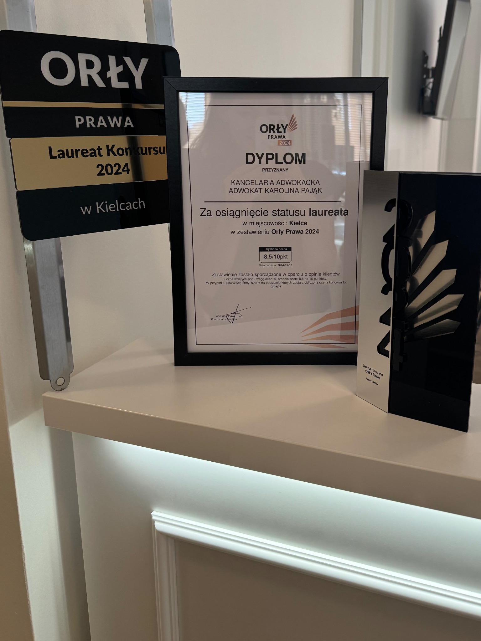 Nagrody i dyplom Kancelarii Adwokackiej Karoliny Pająk, laureata konkursu Orły Prawa 2024 w Kielcach, z oceną 8.5/10, prezentowane na półce.