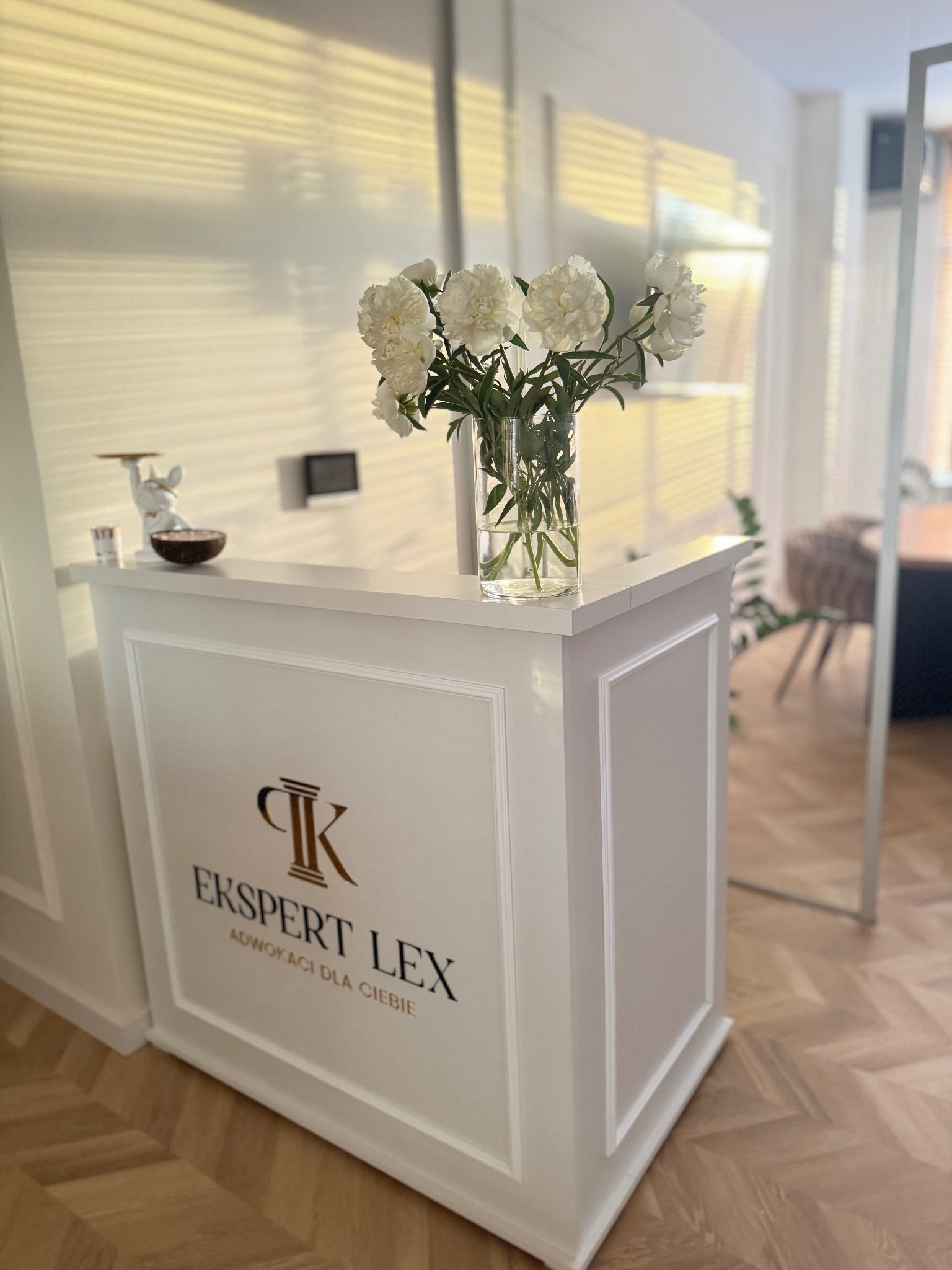 Recepcja w kancelarii prawnej z logo 'Ekspert Lex', bukietem białych piwonii w szklanym wazonie i figurką dekoracyjną na blacie, parkiet w jodełkę na podłodze.