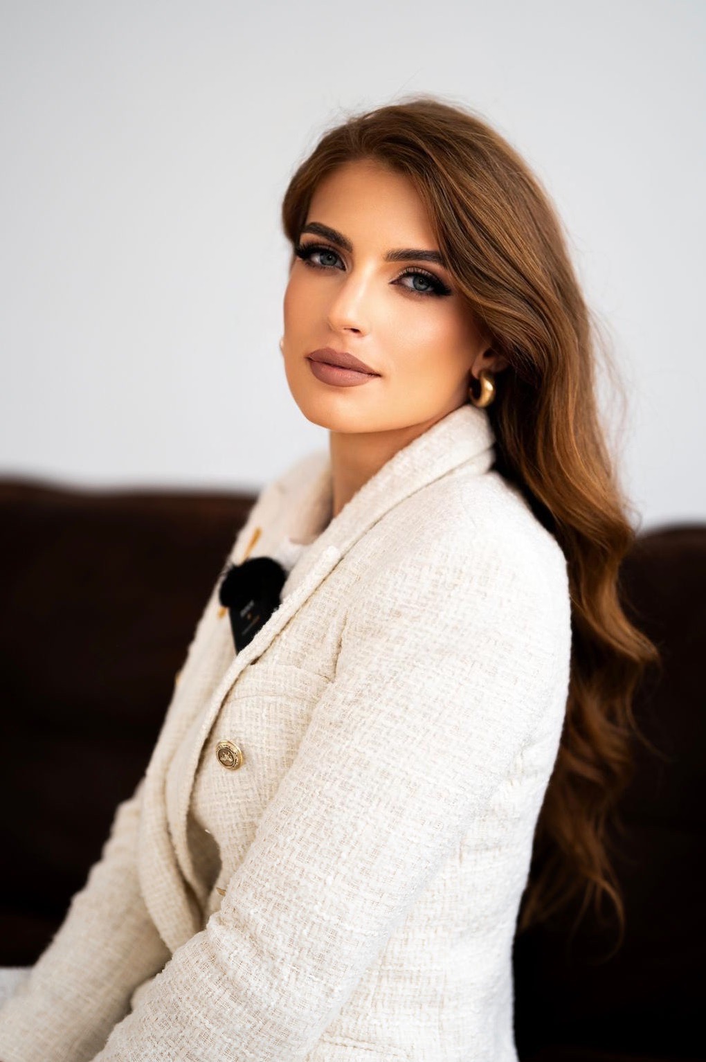 Portret kobiety z długimi brązowymi włosami, ubranej w kremowy żakiet z wyraźną fakturą i złotymi guzikami, siedzącej na ciemnobrązowej sofie.
