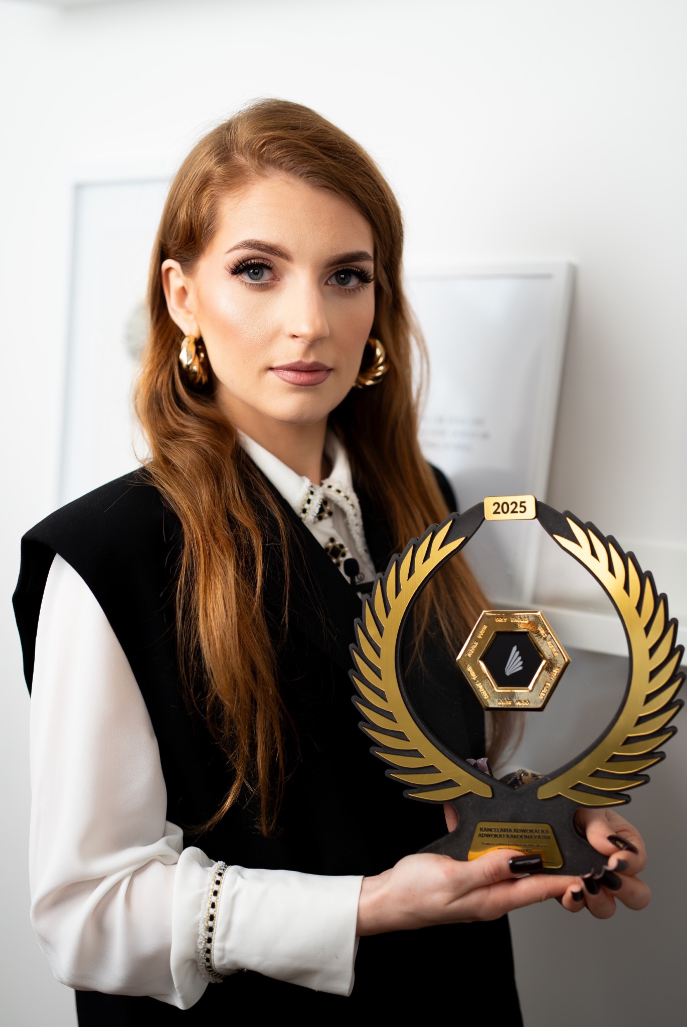 Kobieta z rudymi włosami prezentuje złotą nagrodę prawniczą z napisem 'Kancelaria Adwokacka Adwokat Karolina Pająk 2025' na tle białej ściany.