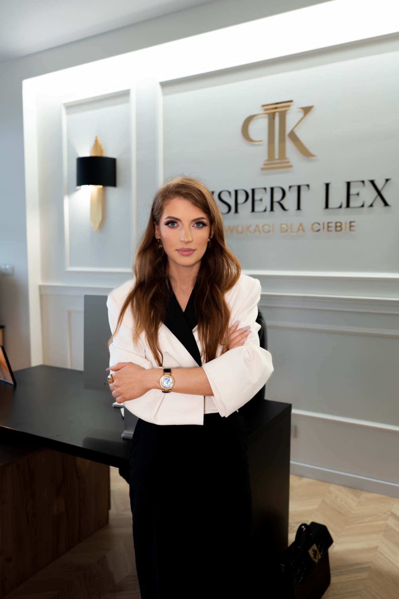 Kobieta w białym żakiecie, skrzyżowane ręce, na tle biura z logo 'Ekspert Lex'. Profesjonalny wizerunek prawnika w nowoczesnym wnętrzu kancelarii.