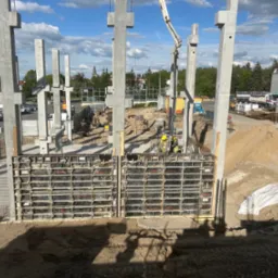 Budowa domu: betonowe słupy konstrukcyjne w szalunkach, widoczni pracownicy w żółtych kaskach, na tle błękitnego nieba z chmurami. Etap wznoszenia budynku.