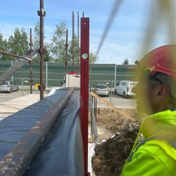 BAR-KAM GROUP SP. Z O.O - Pracownik w kasku i odblaskowej kamizelce używa poziomicy do sprawdzenia pionu konstrukcji szalunkowej fundamentu na placu budowy. Widoczne są elementy zbrojenia.