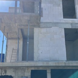 BAR-KAM GROUP SP. Z O.O - Surowy stan dwukondygnacyjnego domu z betonu komórkowego, widoczne rusztowania i drabiny, błękitne niebo w tle. Budowa w toku, brak wykończenia elewacji.