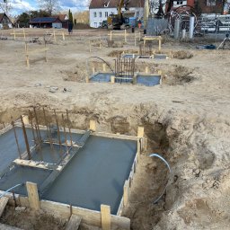 BAR-KAM GROUP SP. Z O.O - Wylane fundamenty z drewnianym szalunkiem i zbrojeniami na placu budowy. W tle budynki mieszkalne i koparka.