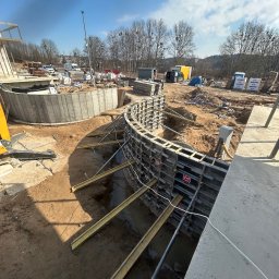 BAR-KAM GROUP SP. Z O.O - Wykop fundamentów z szalunkiem łukowym, wsparty żółtymi belkami. W tle widoczny częściowo zalany betonem fundament i plac budowy z materiałami.